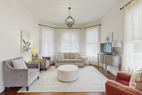 Bright living room with bay windows, beige sofa, purple armchairs, white ottoman, and pendant chandelier overhead.
